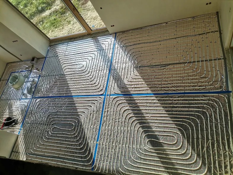 Układana instalacja ogrzewania podłogowego na folii izolacyjnej, widok z góry, rury ułożone spiralnie w kilku sekcjach, siatka montażowa na wierzchu.