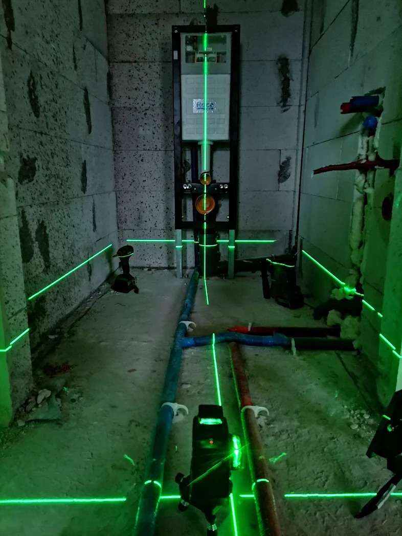 Surowy stan łazienki w trakcie prac hydraulicznych, widoczny stelaż podtynkowy WC z poziomymi i pionowymi liniami lasera wyznaczającymi położenie instalacji, rury wodne i kanalizacyjne w podłodze.