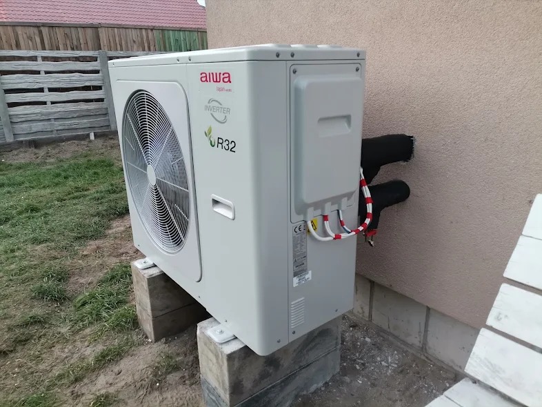 Zewnętrzna jednostka klimatyzacji Aiwa Inverter R32 zamontowana na betonowych bloczkach przy ścianie budynku z widocznymi rurami w izolacji i przewodami elektrycznymi.