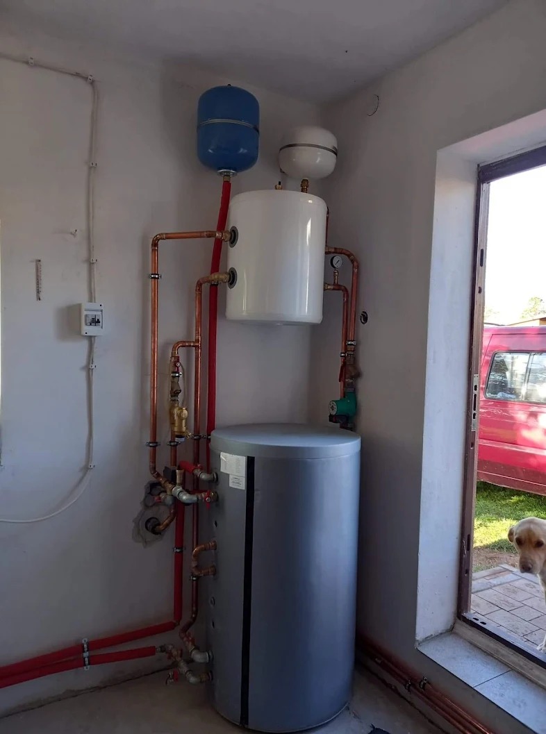 Kompletna instalacja grzewcza z szarym bojlerem, białym zbiornikiem wyrównawczym i niebieskim naczyniem przeponowym, widoczna z perspektywy z dołu, z psem spoglądającym przez drzwi w tle.