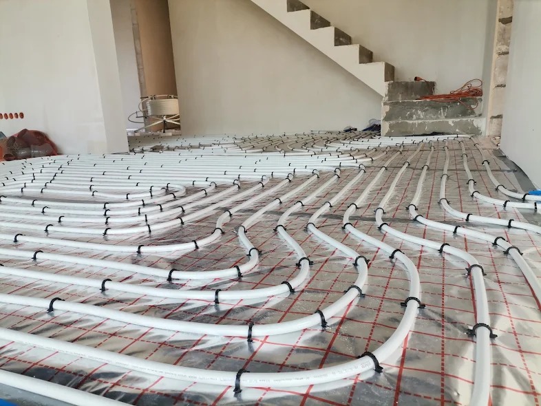 Białe rury ogrzewania podłogowego, równomiernie rozłożone na folii izolacyjnej z czerwoną siatką, widok na surową klatkę schodową w tle.