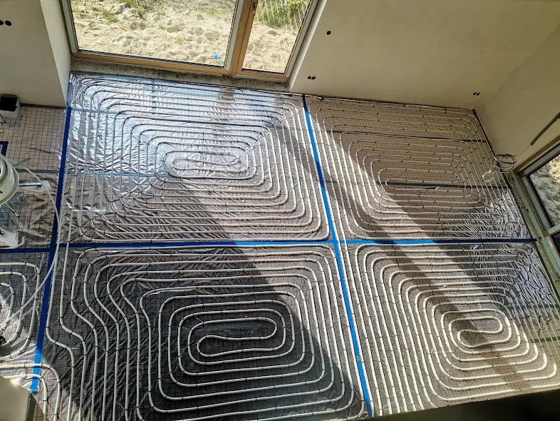 Instalacja ogrzewania podłogowego z rur ułożonych w pętle na folii aluminiowej, widoczne okno dachowe i fragment ściany.