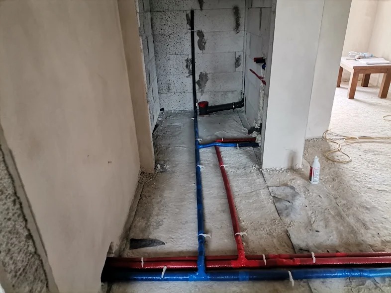 Instalacja wodno-kanalizacyjna w surowym stanie, widoczne rury w kolorze czerwonym i niebieskim, przymocowane do podłoża w nowym budynku, ściany z betonu komórkowego.
