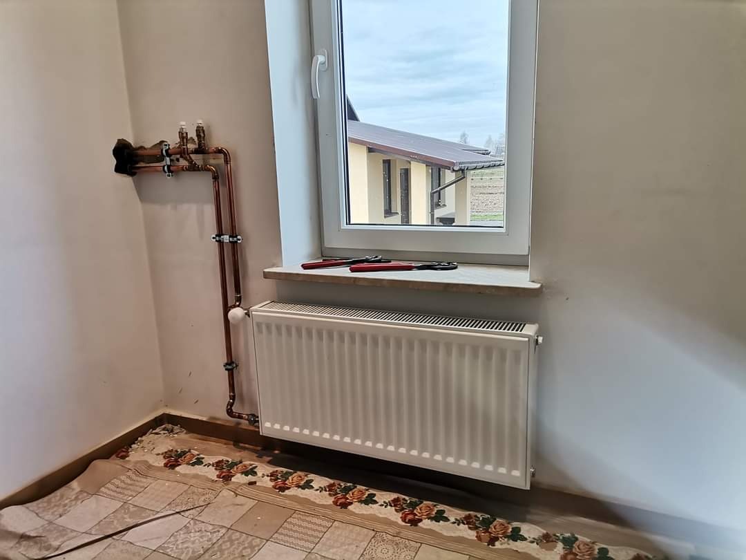 Nowy grzejnik zamontowany pod oknem z rurami miedzianymi, obok leżą szczypce, widok na budynek z okna.