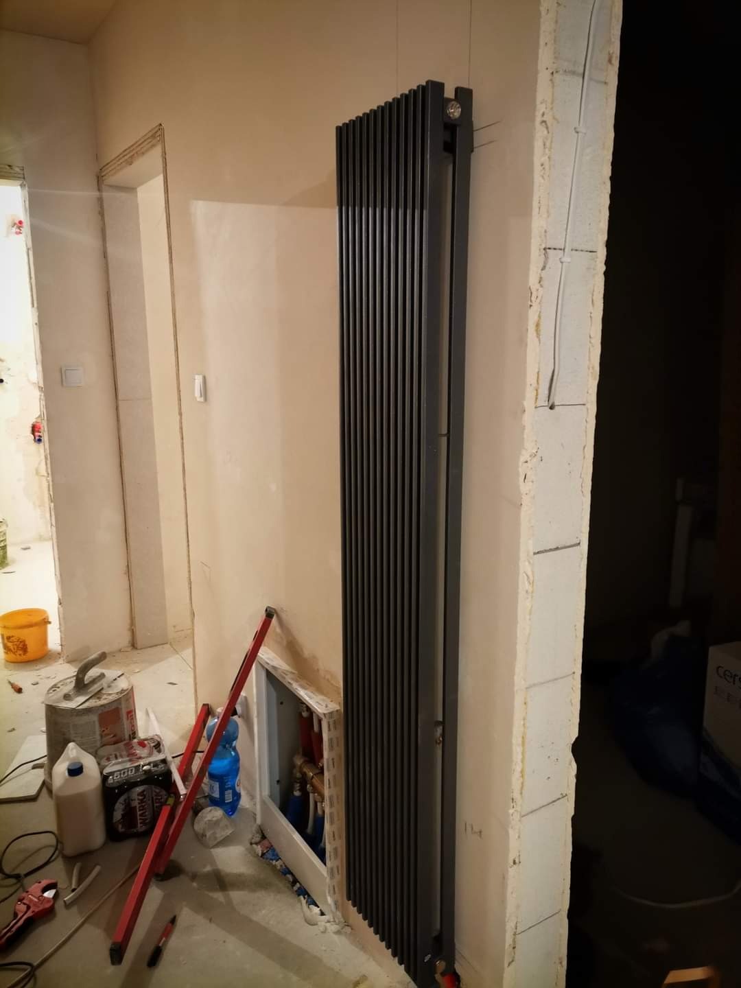 Wysoki, czarny grzejnik panelowy zamontowany na ścianie w trakcie remontu. Widoczne niedokończone obudowy instalacji, narzędzia i materiały budowlane w tle.