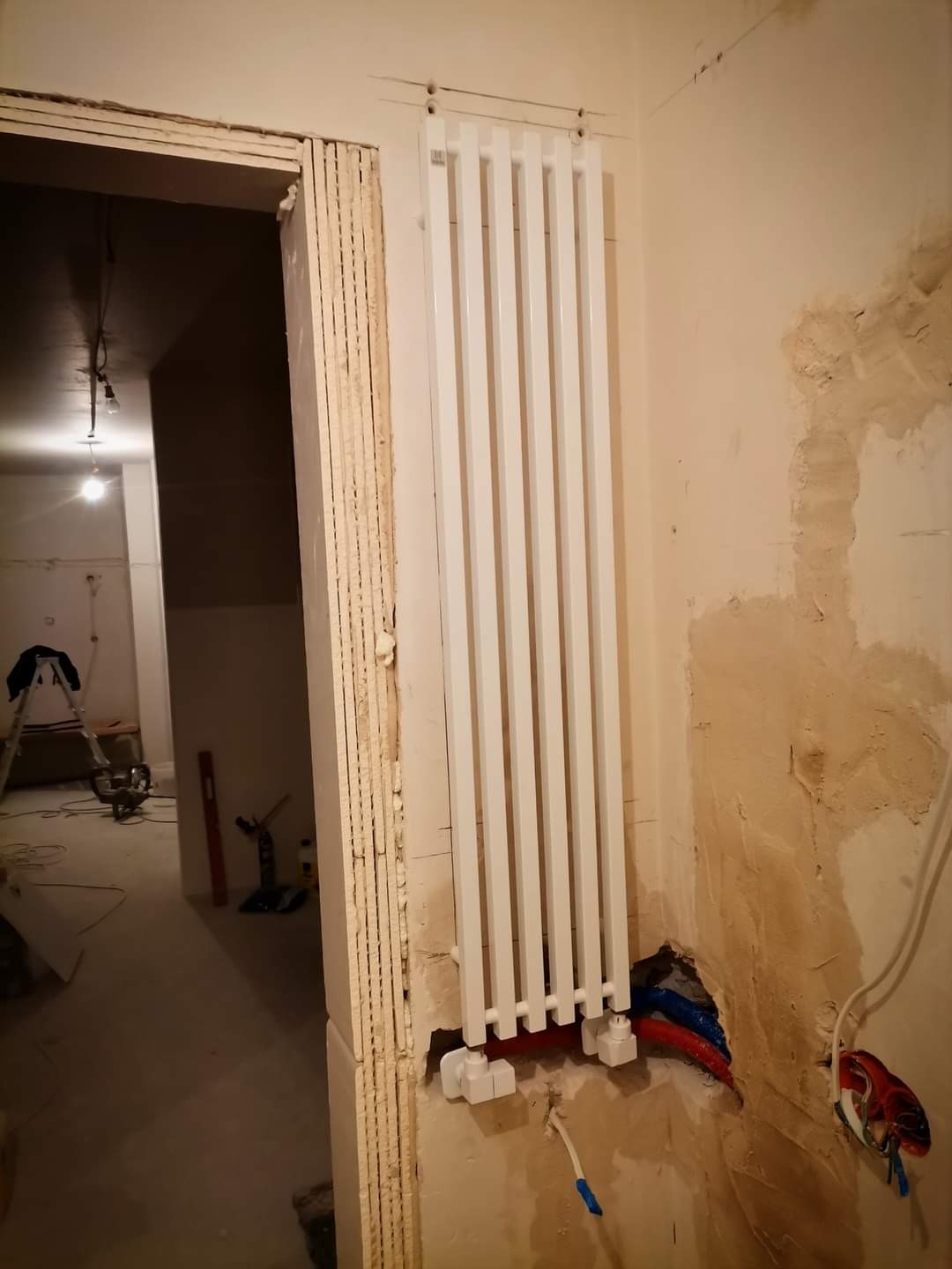 Montaż nowego, białego grzejnika panelowego w surowym wnętrzu. Widoczne rury doprowadzające wodę w kolorach niebieskim i czerwonym oraz niedokończone wykończenie ściany i futryny drzwi.