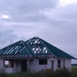 Surowa konstrukcja domu jednorodzinnego z widoczną więźbą dachową, wykonaną z impregnowanego drewna w kolorze zielonym, na tle pochmurnego nieba.