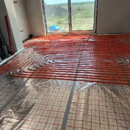 Pomarańczowe rury ogrzewania podłogowego ułożone na folii z siatką w surowym wnętrzu z dużym oknem na ogród.