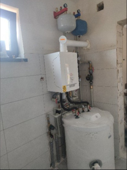 Biały piec gazowy kondensacyjny zamontowany na ścianie w pomieszczeniu z białymi płytkami, widoczne rury, zawory i zbiornik na wodę, instalacja po montażu.