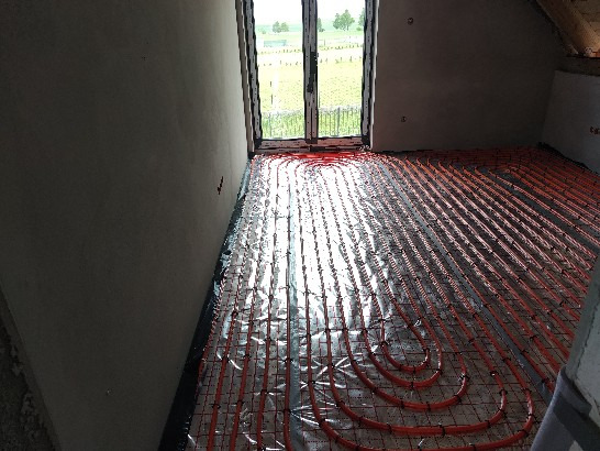 Pomieszczenie w trakcie instalacji ogrzewania podłogowego z widocznymi czerwonymi rurami ułożonymi na folii i przymocowanymi do siatki montażowej, surowe ściany i okno z widokiem na zewnątrz.