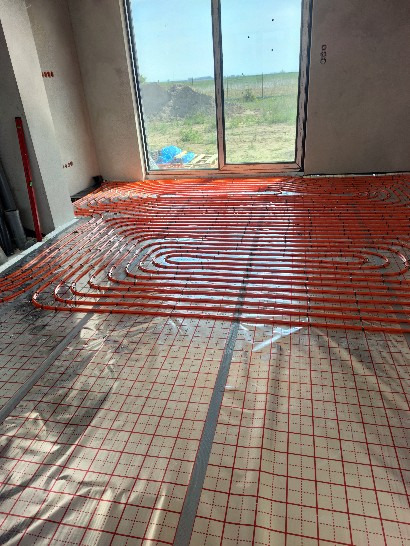 Pomarańczowe rury ogrzewania podłogowego ułożone na folii z siatką w surowym wnętrzu z dużym oknem na ogród.