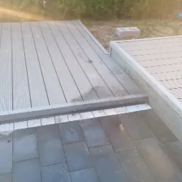 Połączenie szarej deski tarasowej z ciemnymi płytami chodnikowymi, widoczne łączenie dwóch poziomów oraz klimatyzator na ścianie budynku w tle.