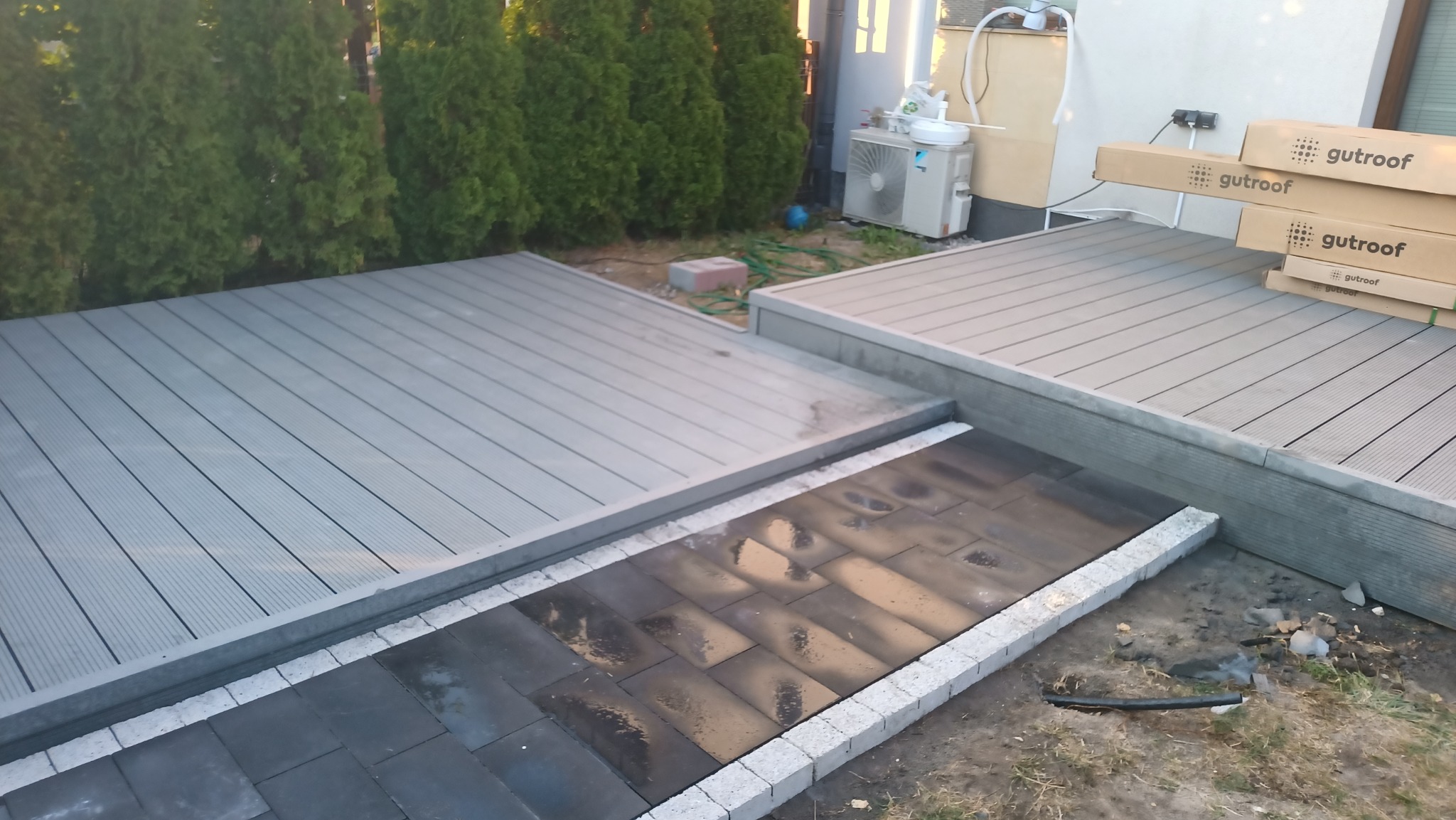 Dwu-poziomowy taras z szarych desek kompozytowych, połączony ścieżką z ciemnej kostki brukowej obramowanej jasnymi obrzeżami, w tle zielone tuje i skrzynie z deskami kompozytowymi.