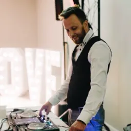 DJ w kamizelce i koszuli, miksujący muzykę na konsoli DJ-ską, w tle podświetlony napis LOVE