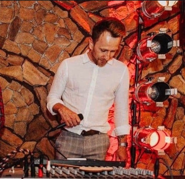 DJ w białej koszuli i kraciastych spodniach, trzymający mikrofon, za nim ściana z kamienia i oświetlenie sceniczne