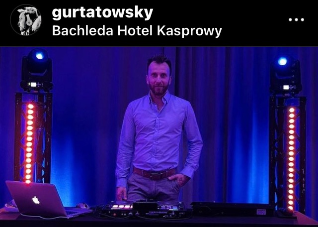 DJ w jasnej koszuli za konsoletą z laptopem, otoczony oświetleniem scenicznym w odcieniach niebieskiego, na tle zasłon.