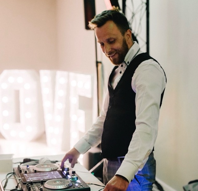 DJ w kamizelce i koszuli, miksujący muzykę na konsoli DJ-ską, w tle podświetlony napis LOVE