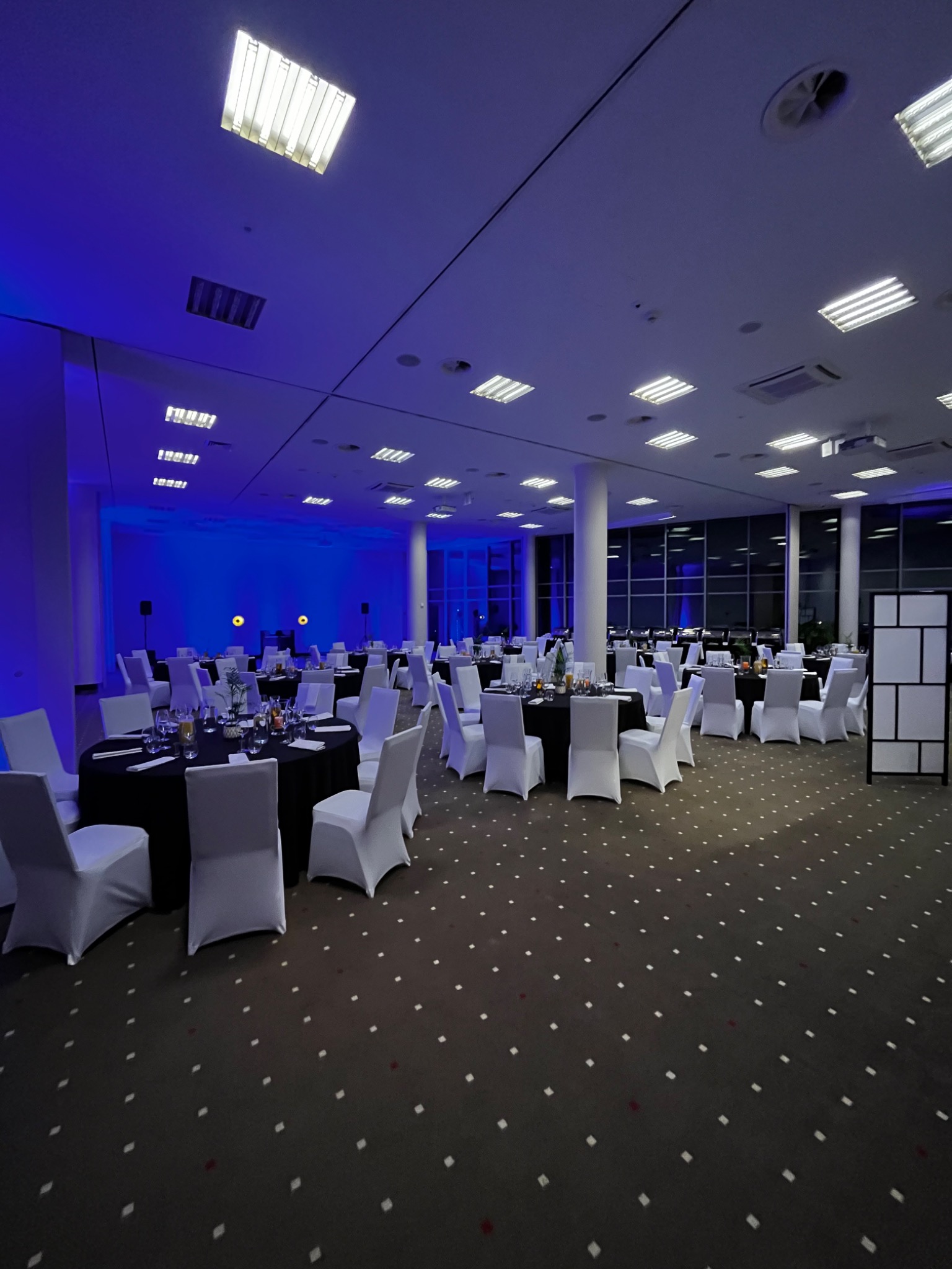 Sala konferencyjna przygotowana na uroczystość, oświetlona niebieskim światłem, z okrągłymi stołami nakrytymi czarnymi obrusami i białymi krzesłami.