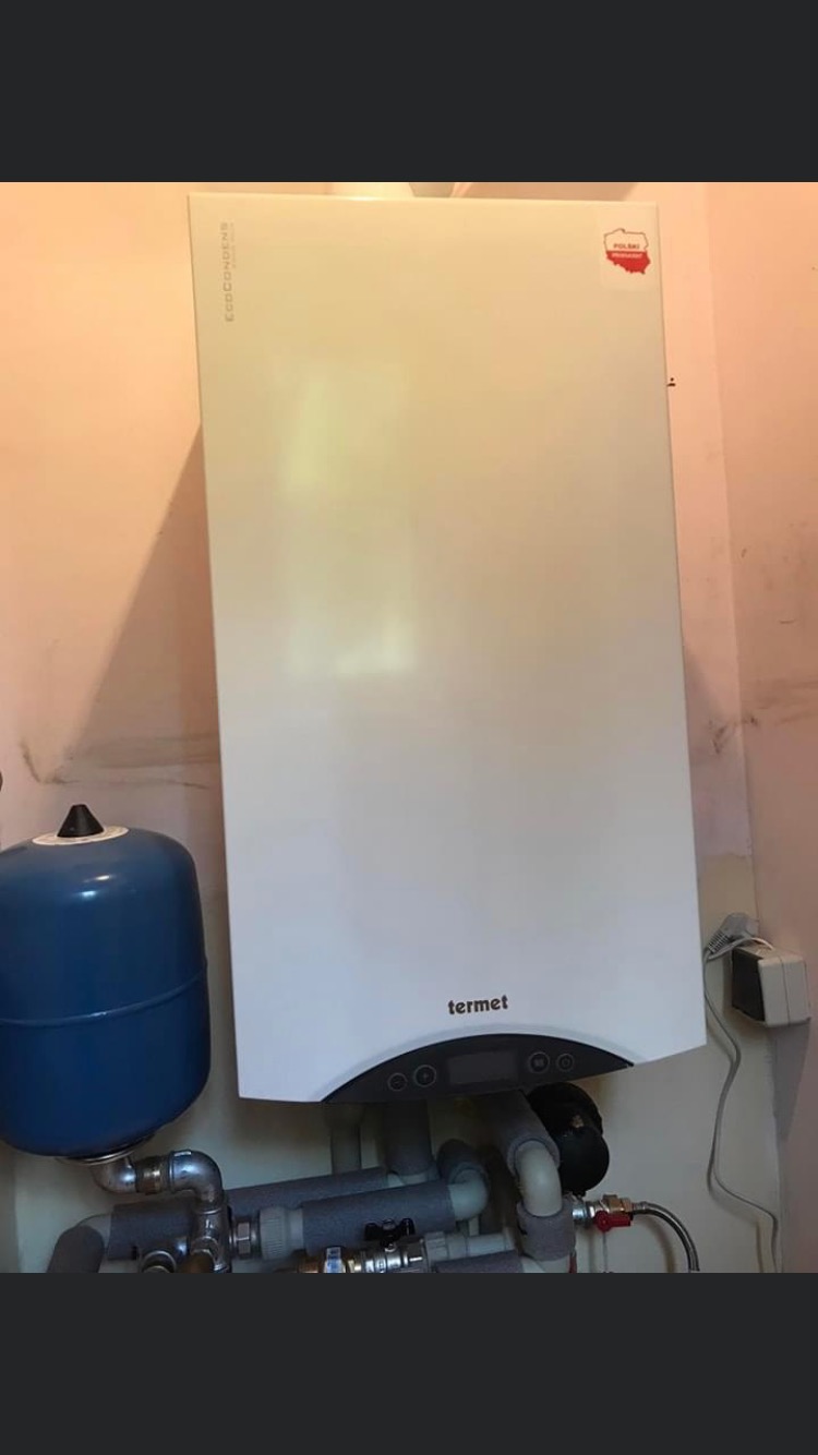 Biały piec gazowy marki Termet zamontowany na ścianie z widocznymi rurami, niebieskim zbiornikiem wyrównawczym i naklejką z konturem Polski.