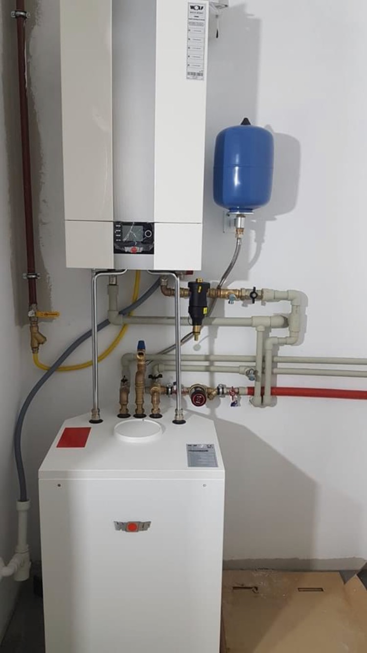 Nowoczesny, dwuczęściowy piec gazowy marki Wolf z podłączonymi instalacjami, w tym niebieski zbiornik wyrównawczy ciśnienia, zamontowany na białej ścianie w nowym pomieszczeniu.