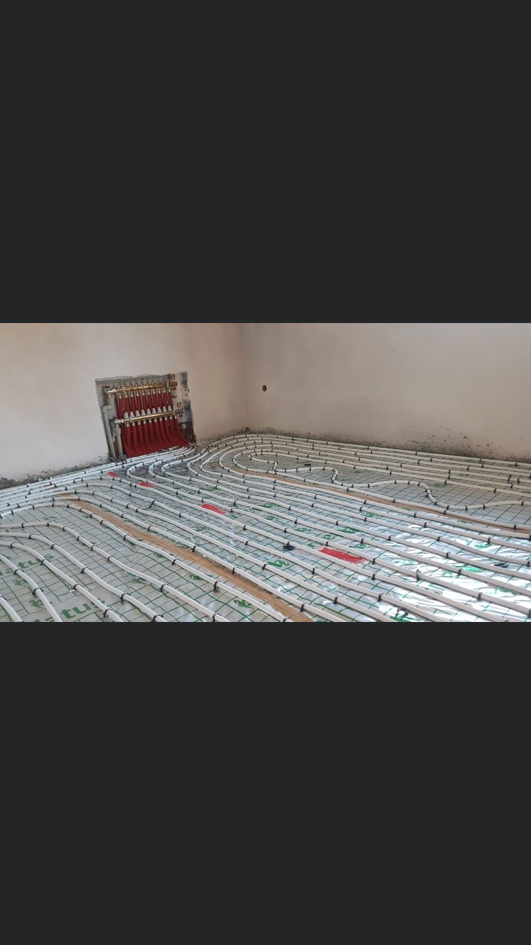 Rozłożone rury ogrzewania podłogowego na macie izolacyjnej, widoczny rozdzielacz z czerwonymi rurami w ścianie, surowy stan wykończenia wnętrza.