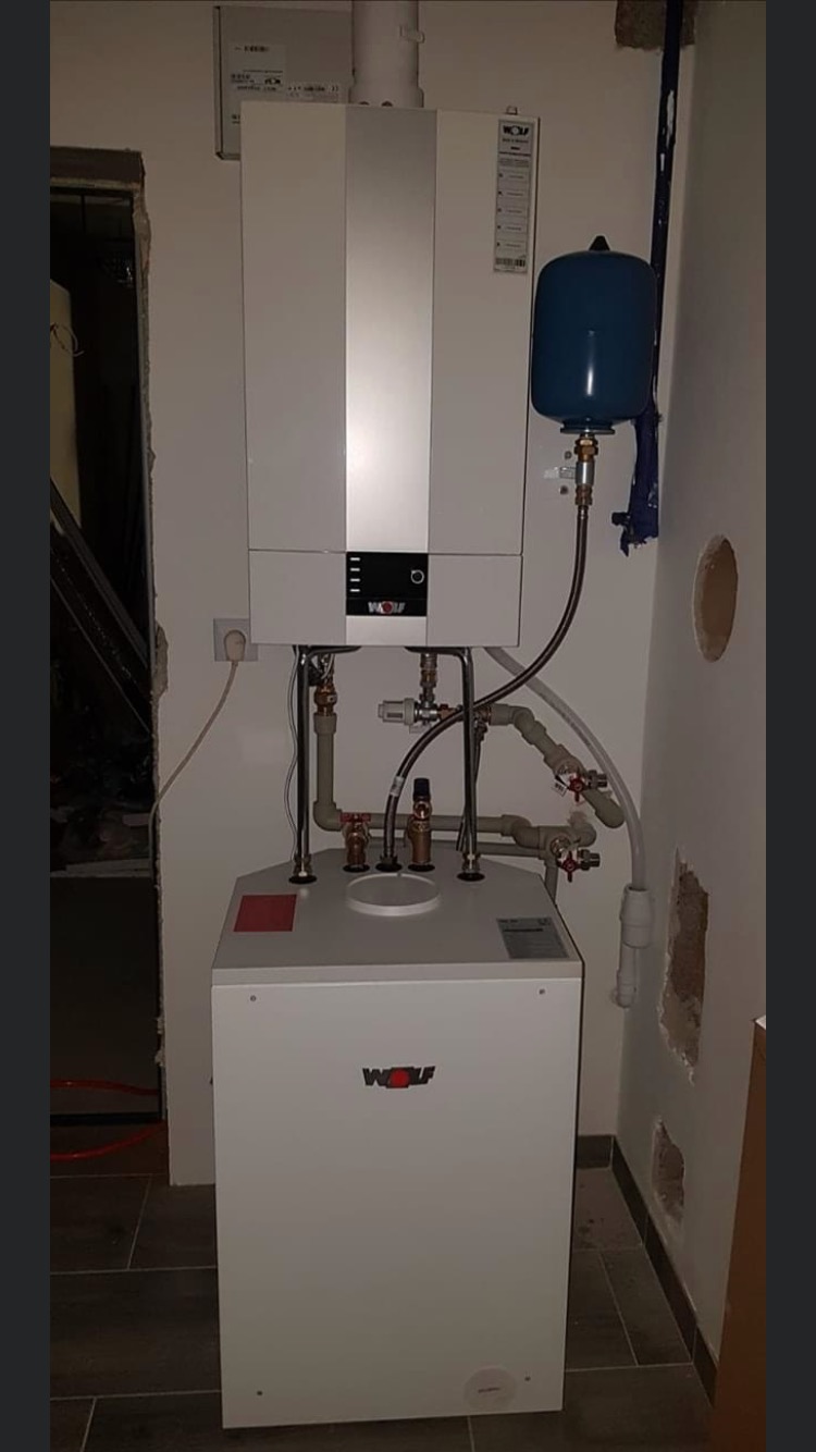 Biały, dwuczęściowy piec gazowy marki Wolf z podłączonymi rurami miedzianymi i zbiornikiem wyrównawczym, zamontowany na szarej podłodze w trakcie remontu pomieszczenia.