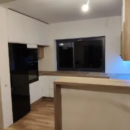 Nowoczesna kuchnia z białymi szafkami, czarną lodówką i blatem drewnianym, oświetlona lampą sufitową i niebieskim światłem pod szafkami wiszącymi.