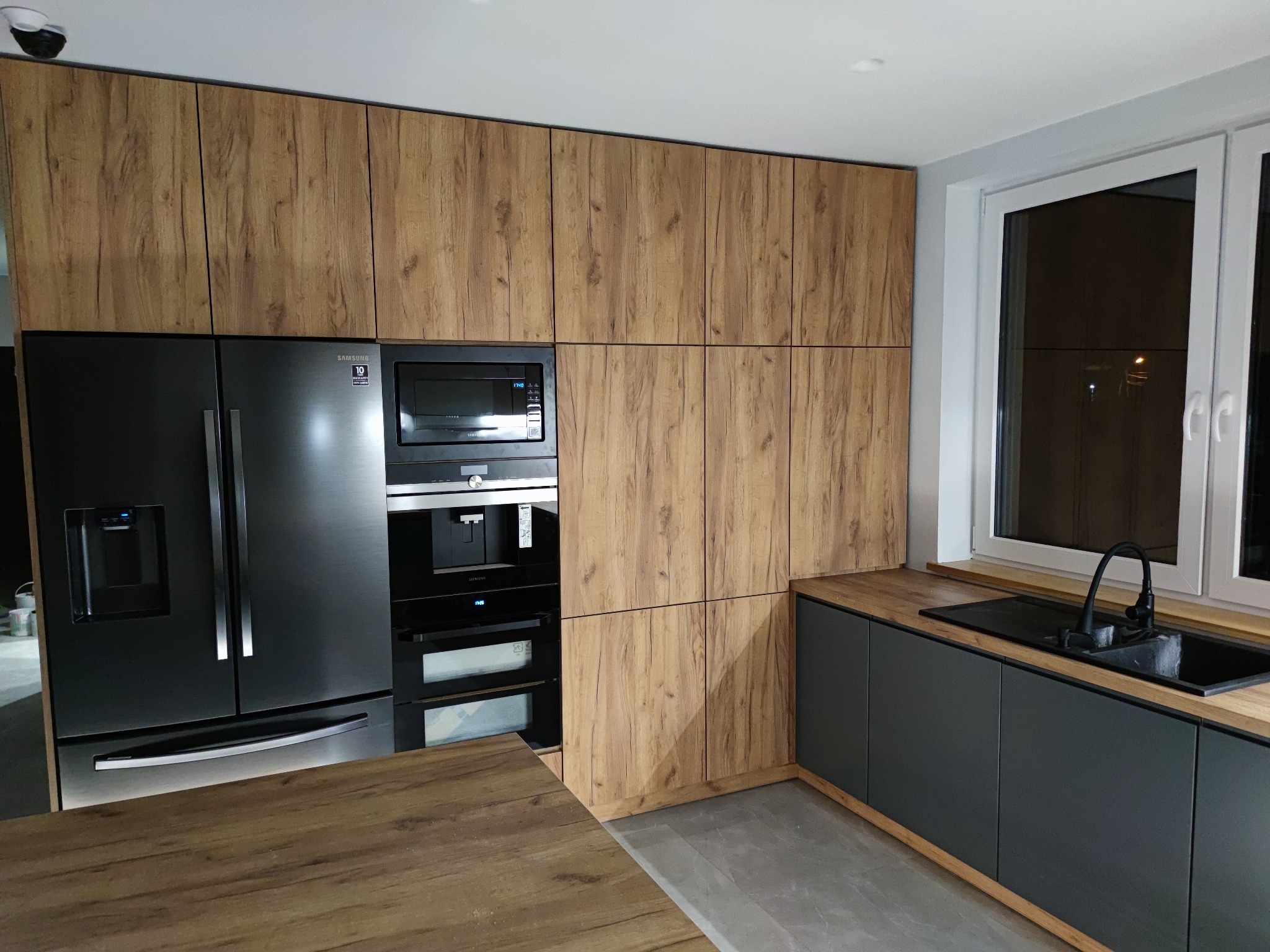 Nowoczesna kuchnia z drewnianymi frontami szafek górnych i czarnymi szafkami dolnymi, czarna lodówka side-by-side i zabudowane urządzenia AGD, widoczny fragment drewnianego blatu.