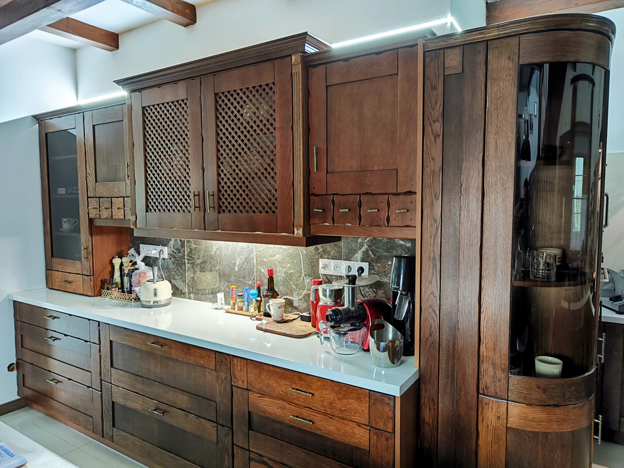 Rustykalna zabudowa kuchenna z ciemnego drewna z frezowanymi frontami i ażurowymi drzwiczkami, biały blat z akcesoriami kuchennymi, oświetlenie podszafkowe LED.