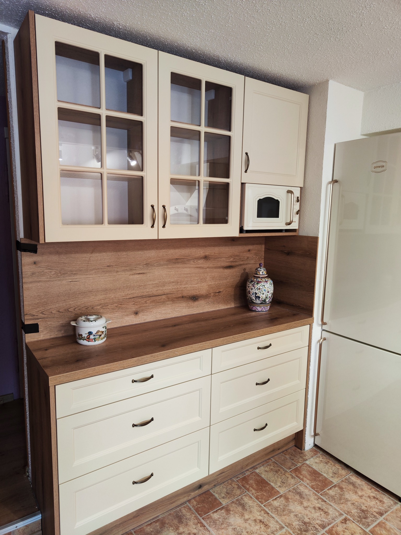 Jasna kuchnia z kremowymi frontami, drewnianym blatem i dekoracyjnymi uchwytami, uzupełniona o szklane witryny górnych szafek z wewnętrznym oświetleniem punktowym, mała mikrofalówka i dekoracyjne...