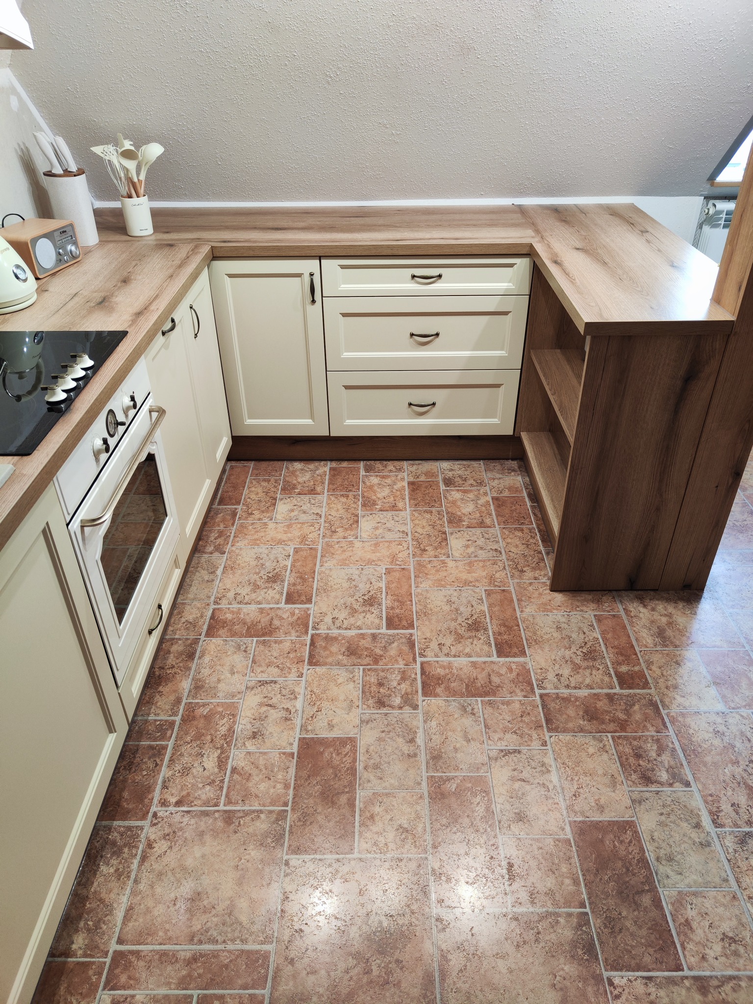 Jasna kuchnia w skosach z drewnianym blatem, beżowymi frontami szafek i brązową podłogą imitującą kamień.