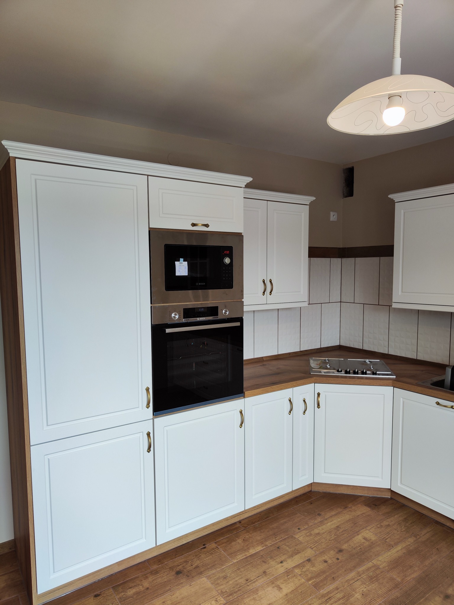 Jasna, klasyczna zabudowa kuchenna z drewnianym blatem i wbudowanym piekarnikiem oraz mikrofalówką, zrealizowana w Kamieńcu Ząbkowickim. Uwagę zwracają frezowane fronty szafek i złote uchwyty.