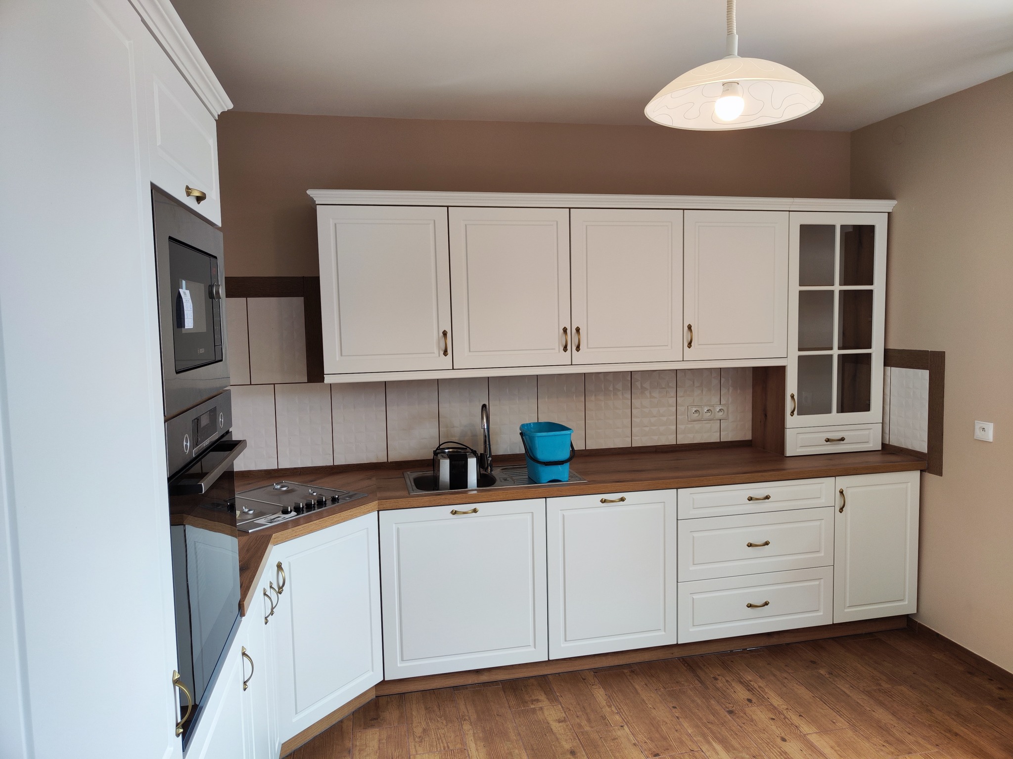 Jasna kuchnia z białymi frontami szafek w stylu klasycznym, brązowy blat, widoczny zlew z akcesoriami i płytą grzewczą, brązowa ściana w tle.