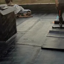 Prace dekarskie na dachu płaskim: dekarz z palnikiem w trakcie zgrzewania papy termozgrzewalnej na tle zachodzącego słońca i pagórkowatego krajobrazu, drugi pracownik robi zdjęcie telefonem.