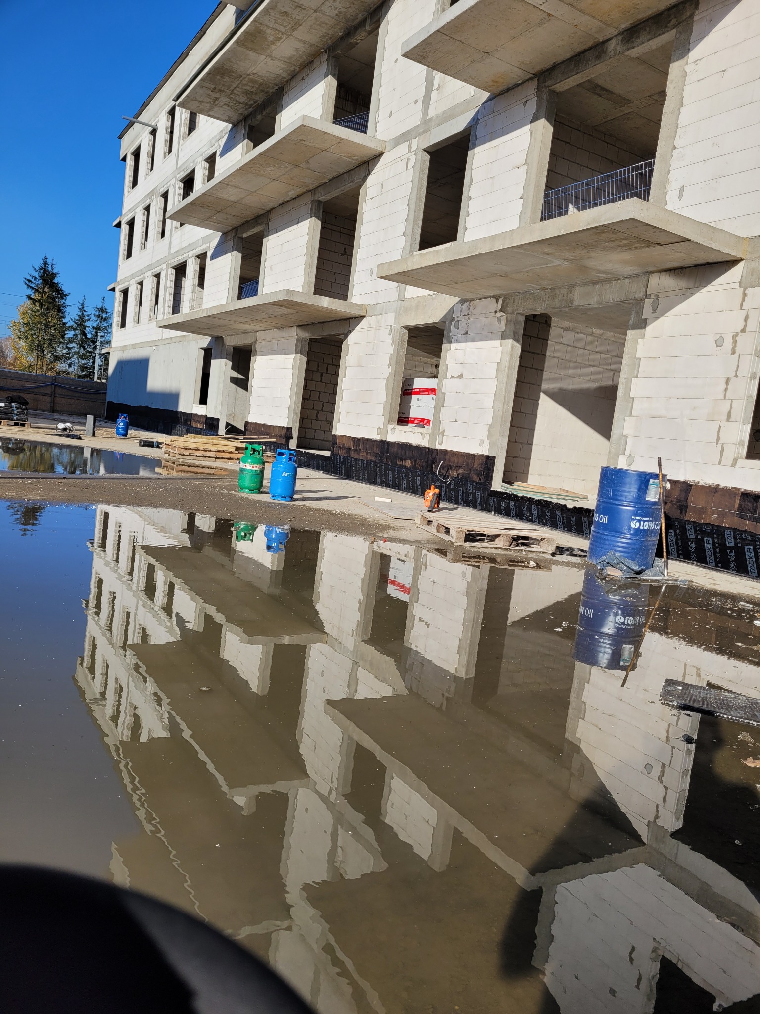 Niedokończony budynek wielopiętrowy z betonu komórkowego odbijający się w dużej kałuży, widoczne butle gazowe i beczka z olejem na placu budowy.