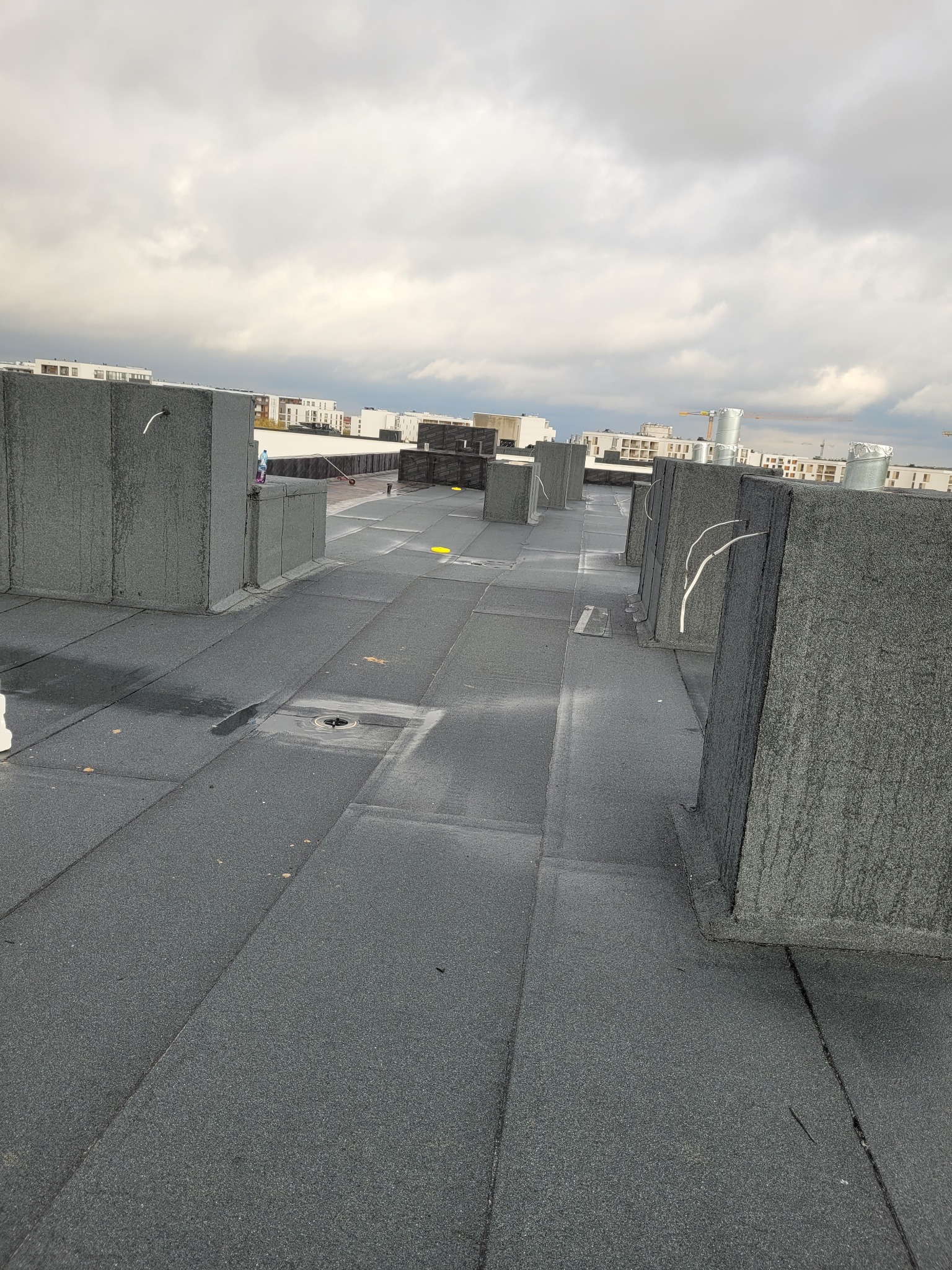 Płaski dach pokryty papą termozgrzewalną z widocznymi kominami wentylacyjnymi i odpływem wody, na tle zabudowa miejska pod pochmurnym niebem.