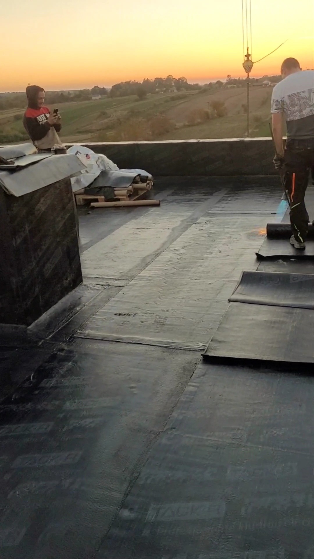 Prace dekarskie na dachu płaskim: dekarz z palnikiem w trakcie zgrzewania papy termozgrzewalnej na tle zachodzącego słońca i pagórkowatego krajobrazu, drugi pracownik robi zdjęcie telefonem.