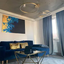 Nowoczesny salon z niebieską, welurową sofą, złotymi poduszkami, szklanym stolikiem kawowym ze złotą podstawą, abstrakcyjnym obrazem na ścianie i okrągłymi, złotymi lampami sufitowymi.