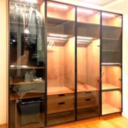 Duża, przeszklona szafa z drewnianymi elementami, oświetleniem LED i czarnymi ramami, widok na ubrania wiszące i leżące na półkach oraz szuflady, podłoga z parkietu.