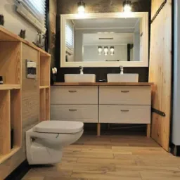 Nowoczesna łazienka z drewnianymi akcentami: drewniana podłoga, szafka pod umywalki z białymi frontami i drewnianym blatem, duże lustro w białej ramie, toaleta podwieszana i drewniane drzwi...