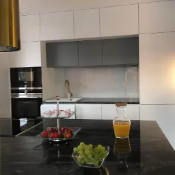 Nowoczesna kuchnia z białymi i szarymi szafkami, czarnym blatem z owocami i sokiem, oraz złotym okapem i baterią.