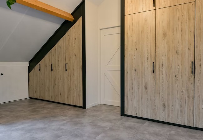 Zabudowa poddasza z drewnopodobnymi frontami szaf, czarne obramowania, szare płytki podłogowe i drewniana belka stropowa.