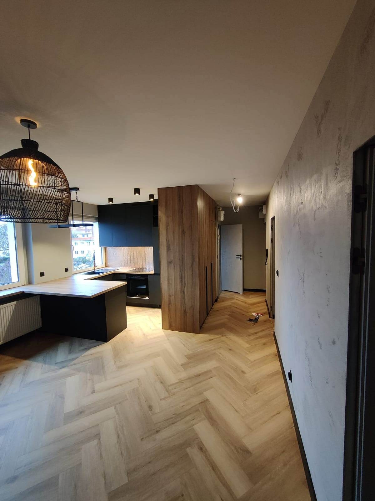 Nowoczesna kuchnia z czarnymi szafkami, drewnianym blatem i szafą w odcieniach drewna, widok na korytarz z podłogą ułożoną w jodełkę.