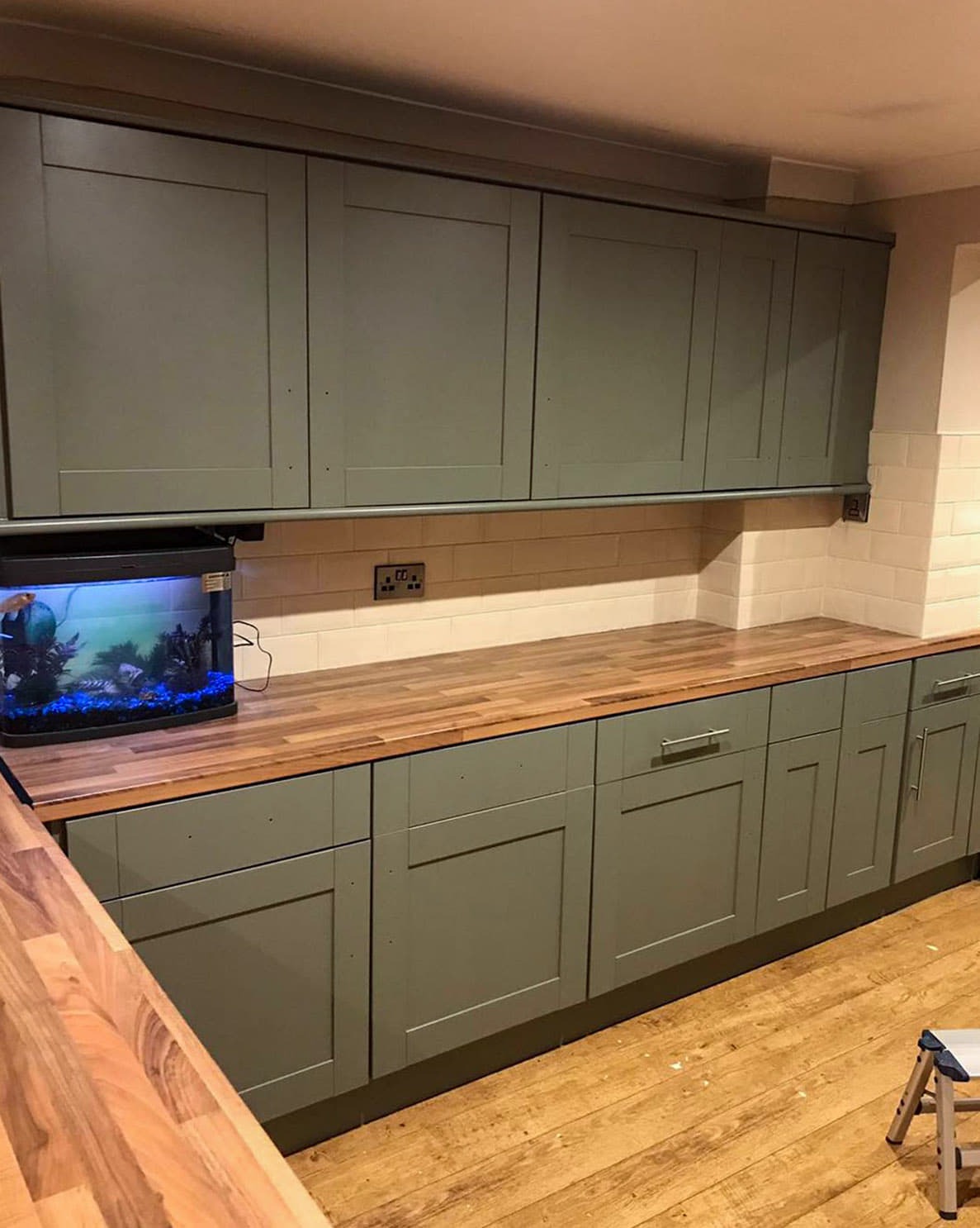 Kuchenne szafki w odcieniach szarości z drewnianym blatem, uzupełnione akwarium z rybkami, widok na fragment podłogi i drabinkę.