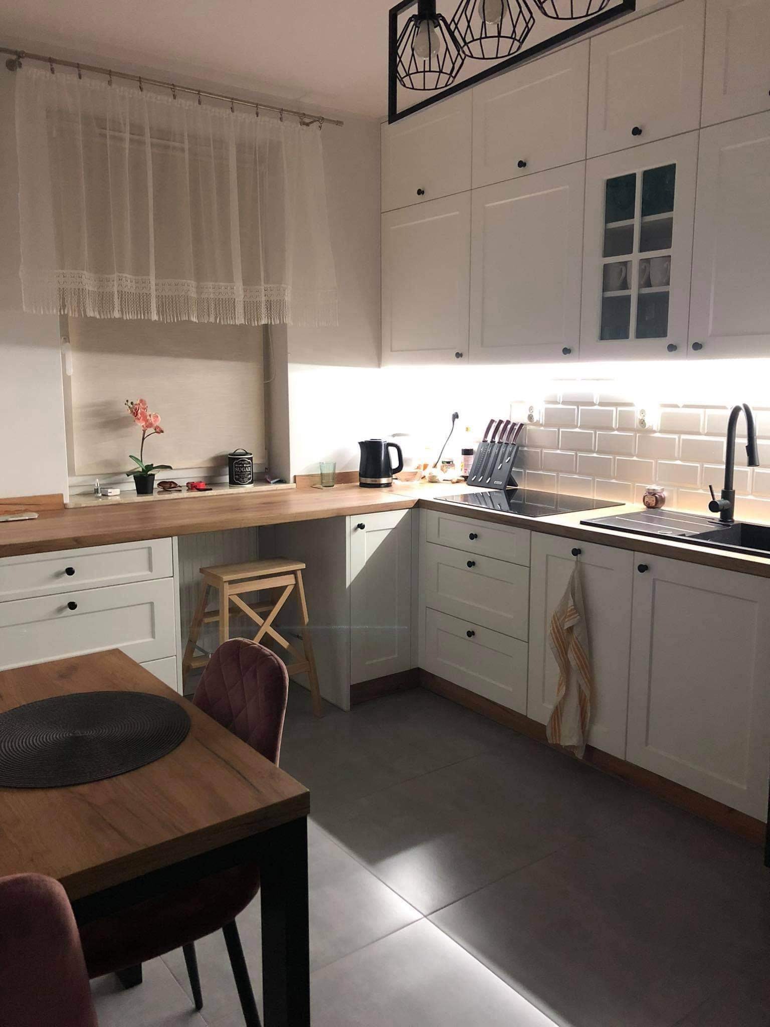 Biała kuchnia z drewnianym blatem, oświetleniem podszafkowym i czarnymi akcentami, takimi jak bateria i uchwyty, uzupełniona drewnianym stołem z czarnymi nogami i różowymi krzesłami.