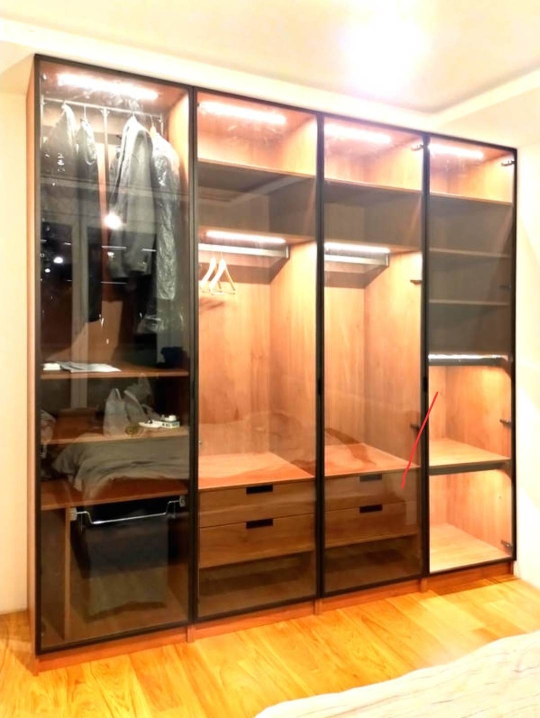Duża, przeszklona szafa z drewnianymi elementami, oświetleniem LED i czarnymi ramami, widok na ubrania wiszące i leżące na półkach oraz szuflady, podłoga z parkietu.