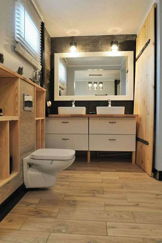 Nowoczesna łazienka z drewnianymi akcentami: drewniana podłoga, szafka pod umywalki z białymi frontami i drewnianym blatem, duże lustro w białej ramie, toaleta podwieszana i drewniane drzwi...