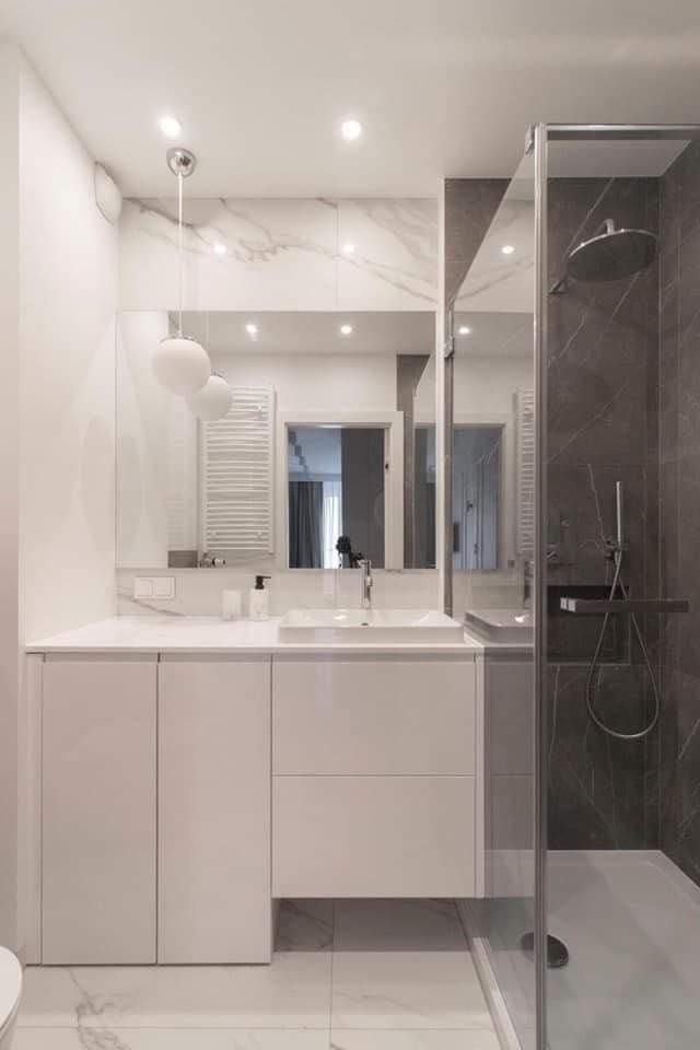 Nowoczesna łazienka z białą szafką pod umywalką, marmurowym blatem i ścianą, szklaną kabiną prysznicową z szarymi płytkami imitującymi marmur oraz kulistymi lampami wiszącymi.