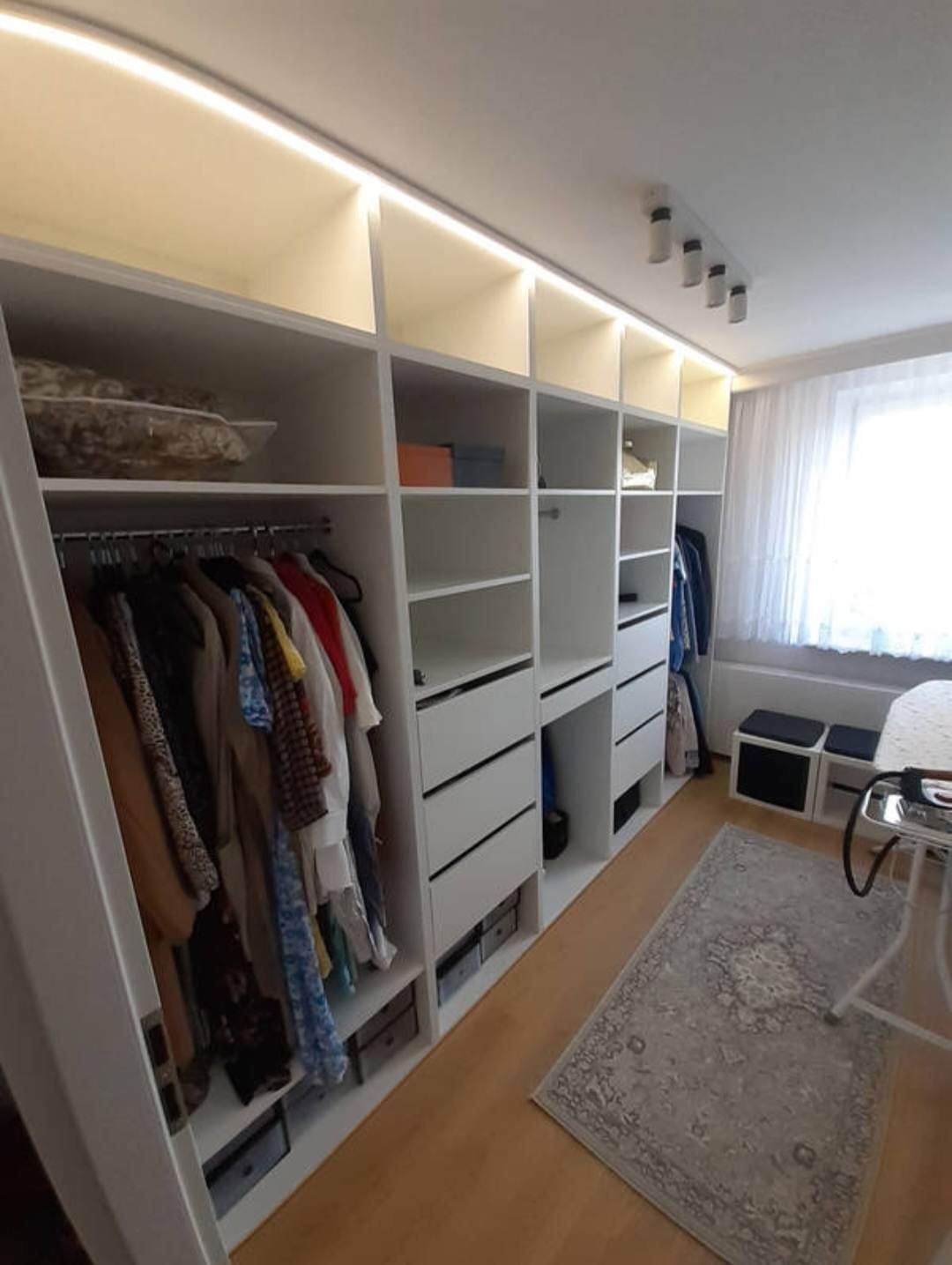 Biała, przestronna garderoba z oświetleniem LED w górnej części, wyposażona w półki, szuflady i drążki na ubrania. Widoczny dywan na podłodze i fragment okna z zasłonami w tle.