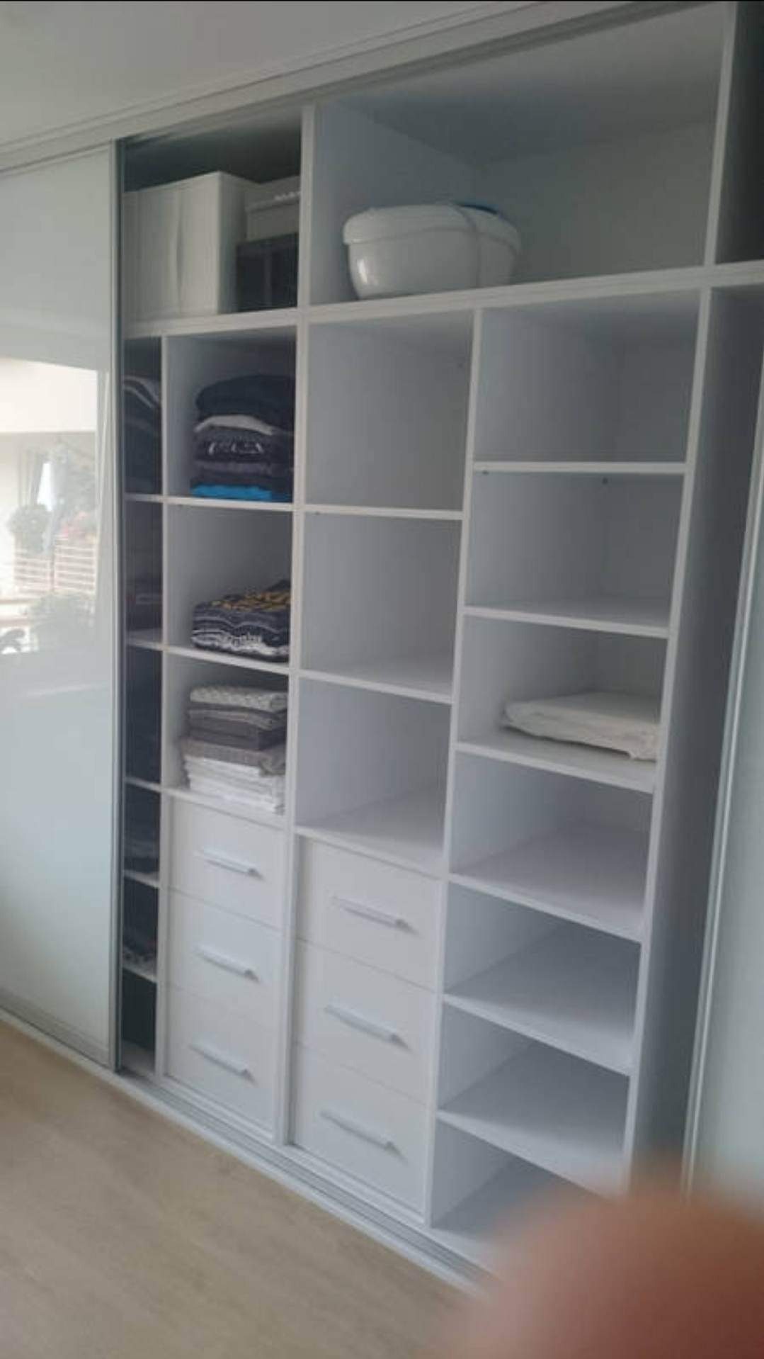 Biała szafa przesuwna z półkami i szufladami, częściowo wypełniona ubraniami i pudełkami, widok z boku.